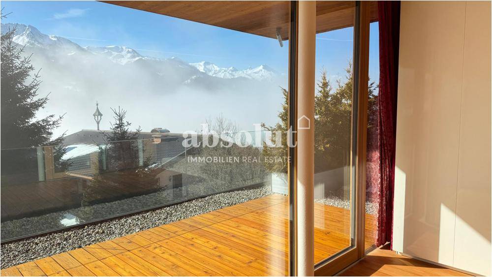 Balkon mit Glasbrüstung und weitem Blick auf die verschneite Berglandschaft und umliegende Häuser.