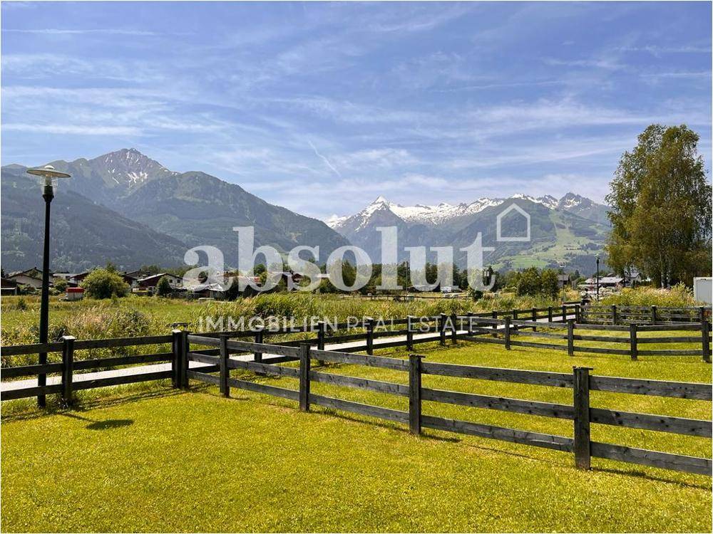 Weitläufige grüne Landschaft mit Holzzäunen und beeindruckendem Bergblick im Hintergrund unter blauem Himmel.