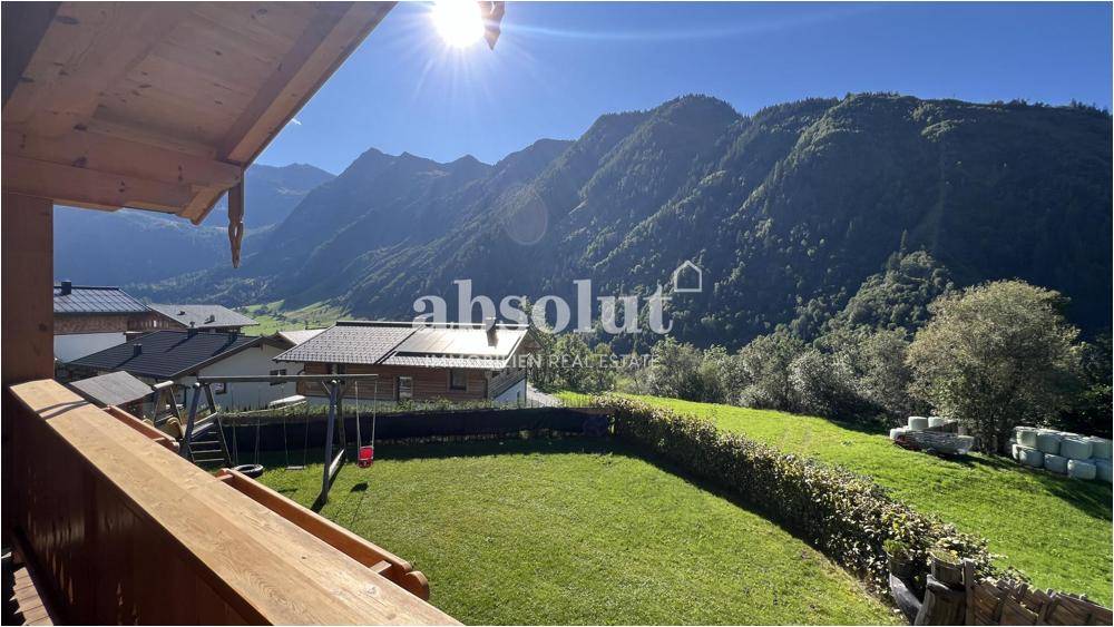 Sonniger Gartenbereich mit Rasenfläche, Hecke und Spielgerät, umgeben von einer beeindruckenden Berglandschaft.