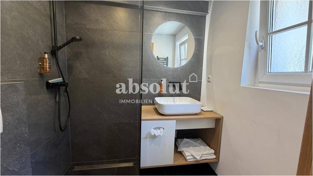 Modernes Badezimmer mit dunklen Fliesen, begehbarer Dusche und einem runden Spiegel.