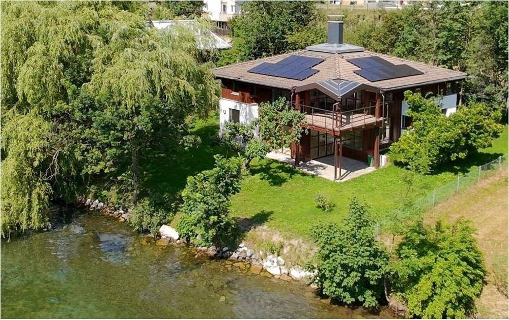 Haus am Seeufer mit Solarpaneelen und großzügigen Terrassenbereichen, umgeben von Grün.