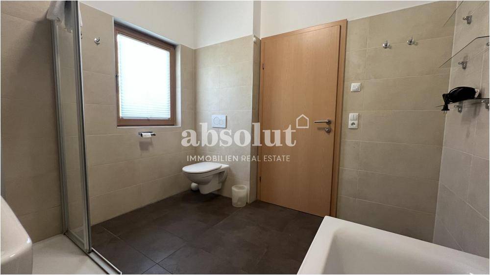 Badezimmer mit Badewanne, WC und Fenster, hellen Fliesen und Holztür.