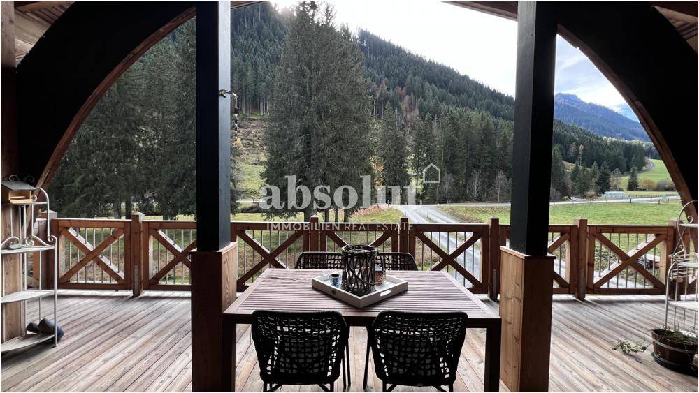 Überdachte Terrasse mit Holzboden und Sitzgelegenheiten, umgeben von grüner Natur und Bergen.