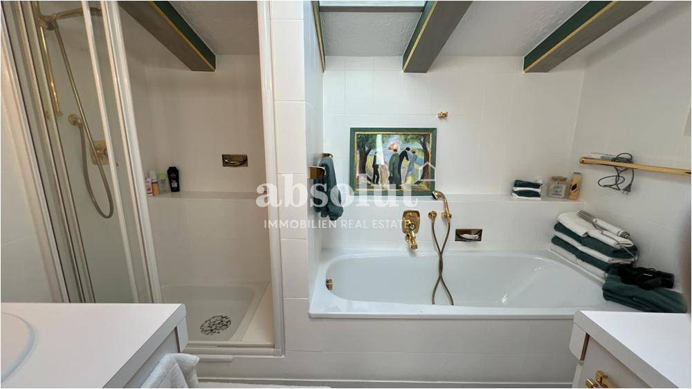 Helles Badezimmer mit Badewanne, separater Dusche und goldenen Armaturen, unter Holzbalken.