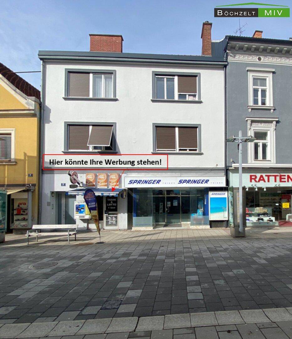 Mehrstöckiges Geschäftsgebäude an einer belebten Straße mit Schaufenstern und Rollläden an den oberen Etagen.