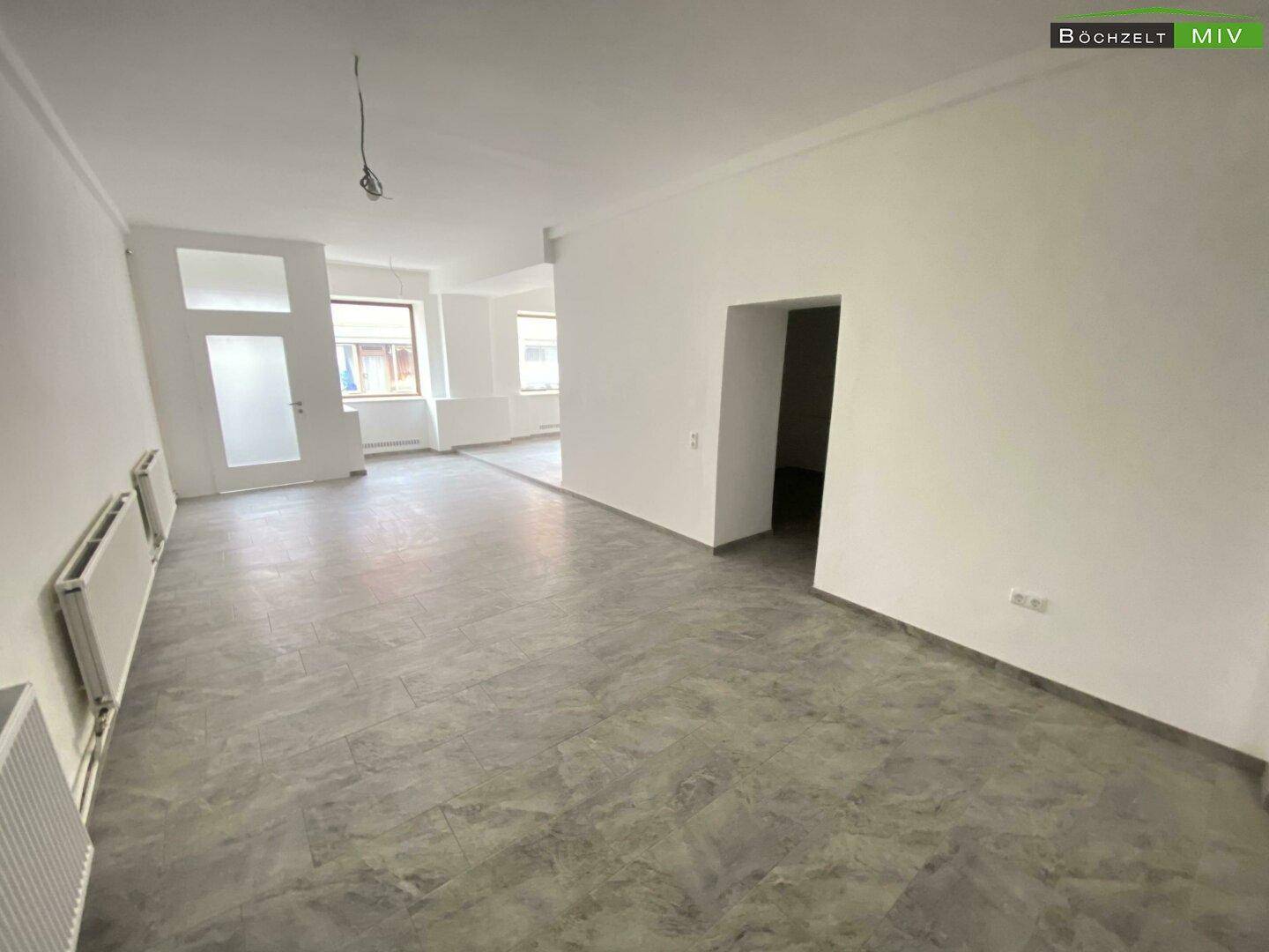Heller, offener Gewerberaum mit grauem Fliesenboden, mehreren Heizkörpern und Blick auf die Stadt.