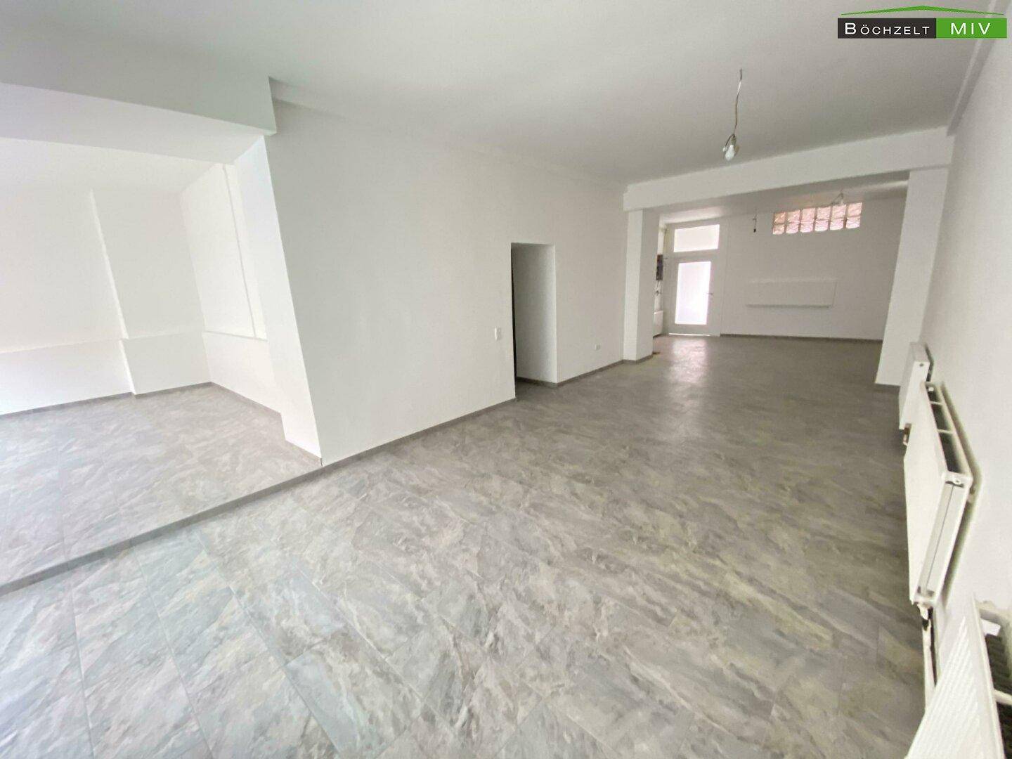 Großer, leerer Gewerberaum mit grauem Fliesenboden, hellen Wänden und Zugang zu weiteren Bereichen.