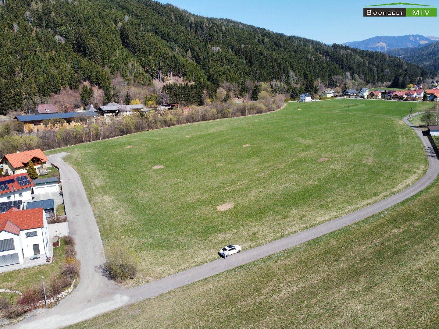 Weitwinkelaufnahme eines grünen Feldes mit Häusern und einer beeindruckenden Bergkette.