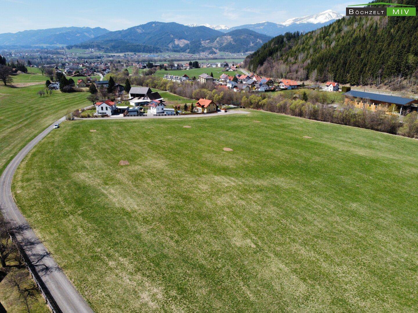 Luftaufnahme eines großen grünen Feldes mit Häusern und Bergen im Hintergrund.