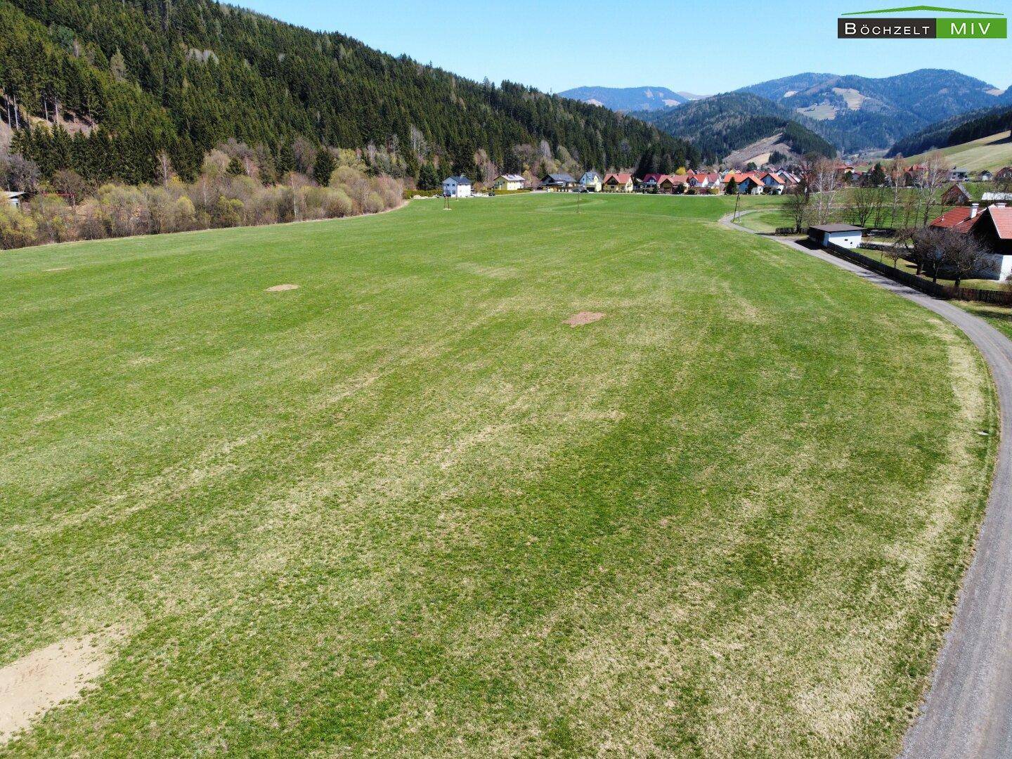 Ausgedehntes grünes Feld mit Blick auf eine ländliche Siedlung und Berge.