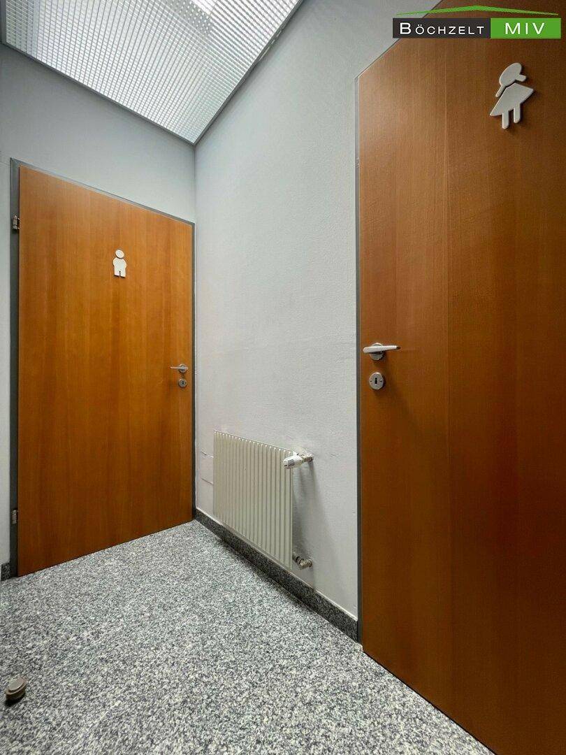 Eingangsbereich zu den Toiletten mit zwei Holztüren, Geschlechter-Symbolen und Fliesenboden.