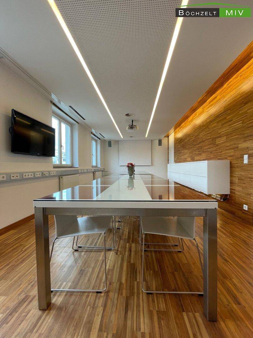 Moderner Konferenzraum mit langem Glastisch, Stühlen, Holzvertäfelung und eleganter Deckenbeleuchtung.