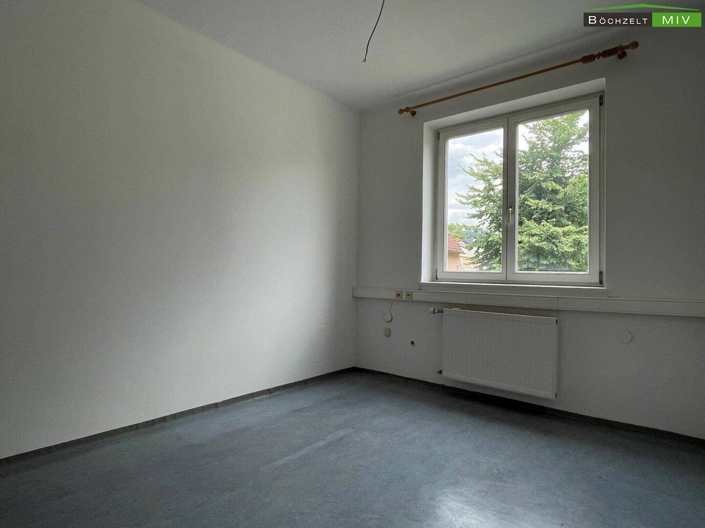 Leerer Raum mit weiß gestrichenen Wänden und einem großen Fenster mit Blick auf Bäume im Freien.
