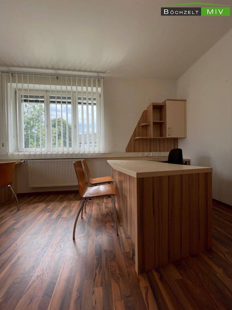 Büroraum mit Holzboden, einem Schreibtisch und Stühlen, sowie einem Fenster mit Jalousien für Privatsphäre.