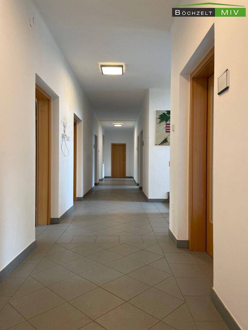 Langer Flur mit grauem Fliesenboden und mehreren Holztüren, beleuchtet von modernen Deckenlampen.