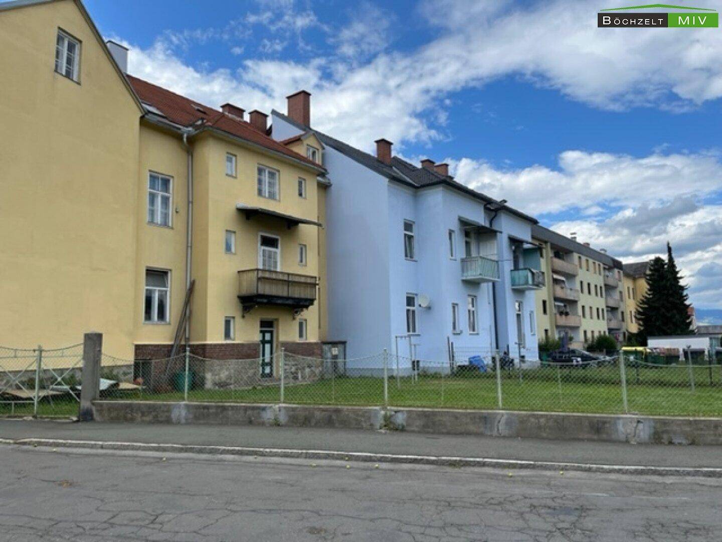 Rückseite der Gebäude mit Balkonen und einer Grünfläche, umgeben von einem Zaun.