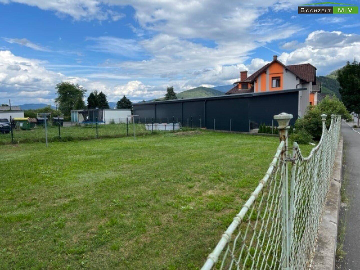 Weitläufige Grünfläche mit Zaun und Blick auf weitere Gebäude und Hügel in der Ferne.