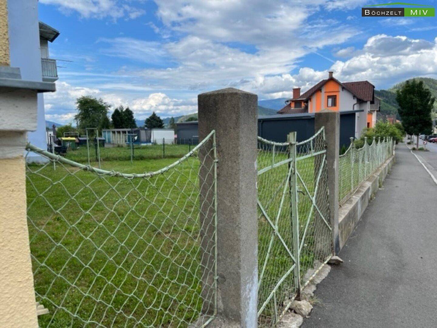 Alter Maschendrahtzaun begrenzt eine grüne Wiese mit Blick auf ein entferntes Haus.