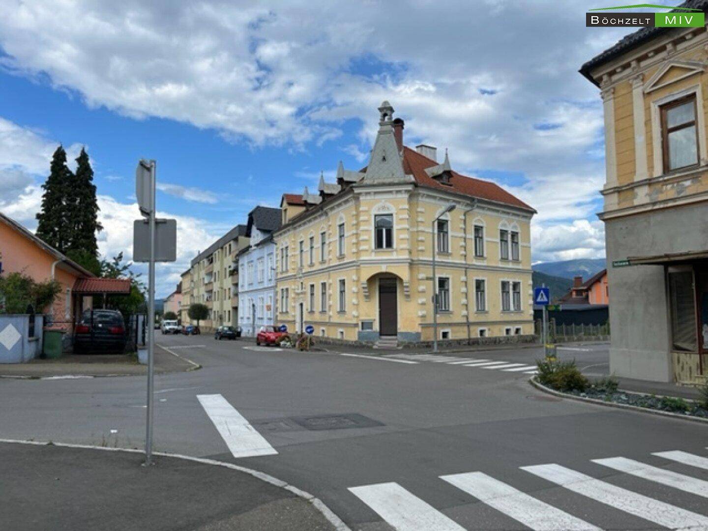 Historisches Gebäude an einer Straßenecke mit umliegenden Häusern und bergiger Landschaft.