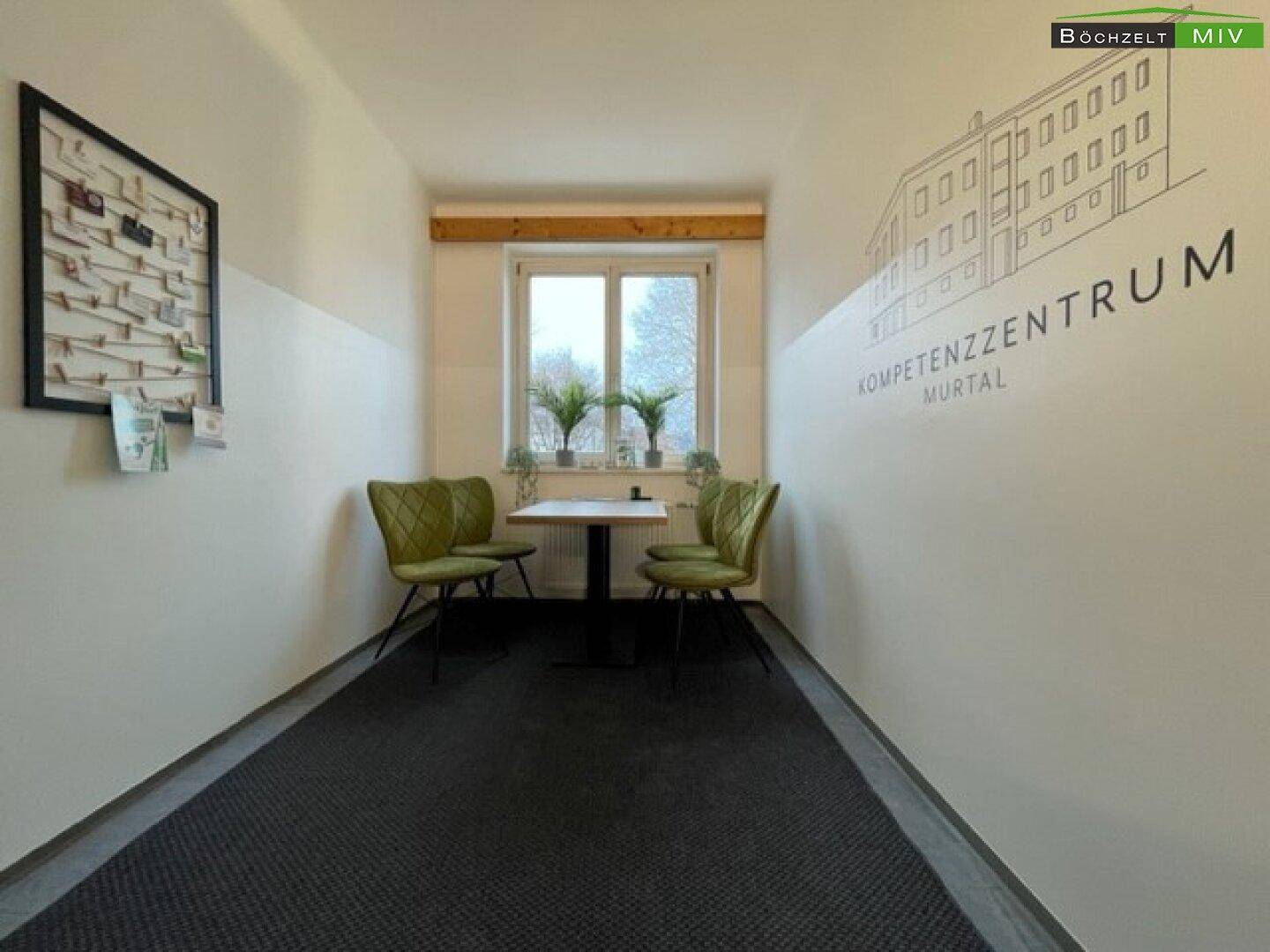 Heller Gemeinschaftsbereich mit Sitzgelegenheiten, einem Tisch und einem Fenster, das Tageslicht hereinlässt.