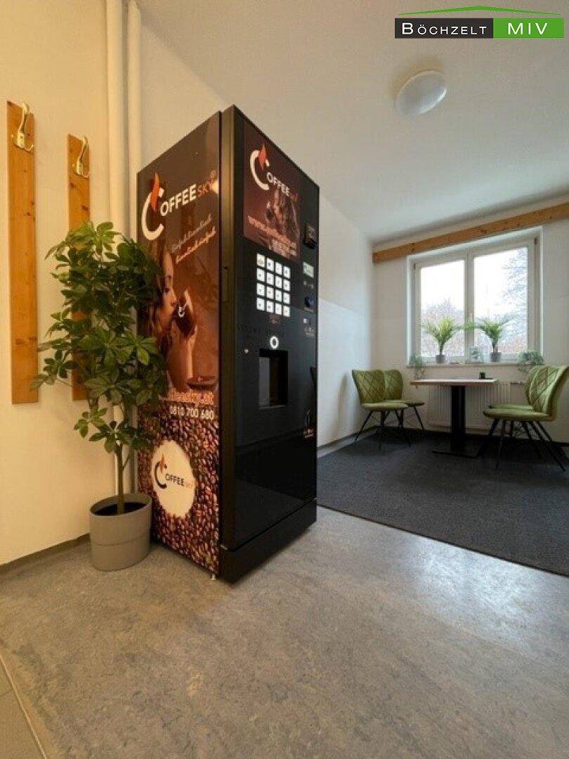 Heller Flurbereich mit einem Kaffeeautomaten, Pflanzen und einem kleinen Sitzbereich im Hintergrund.