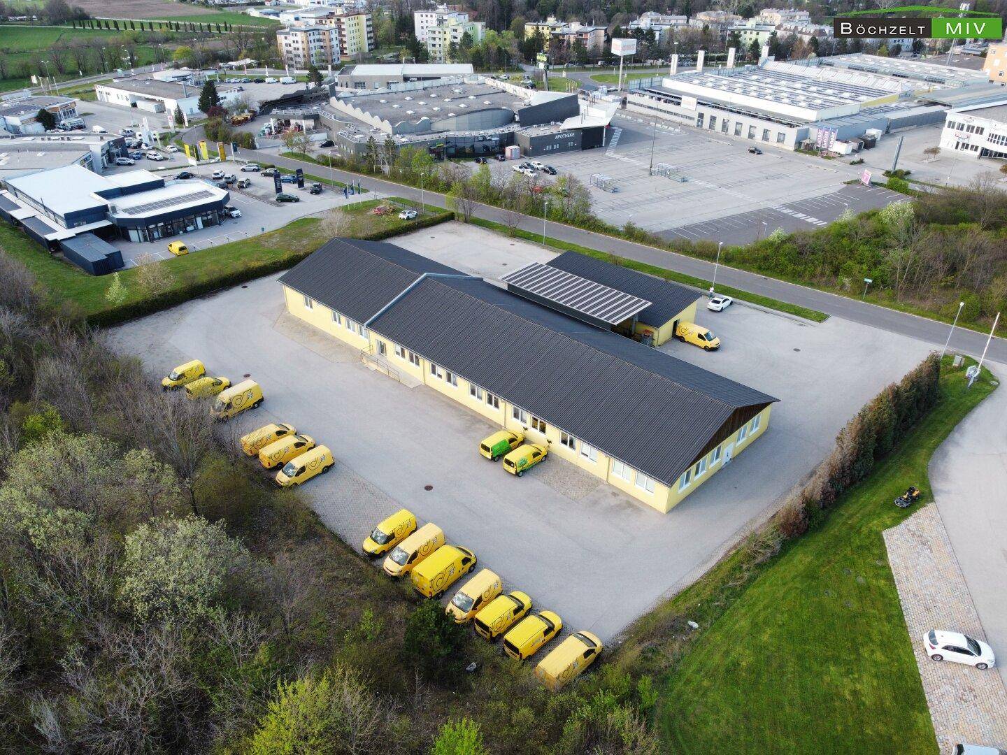 Luftaufnahme eines Gewerbegebäudes mit Parkplätzen und angrenzenden Industriegebäuden im Hintergrund.