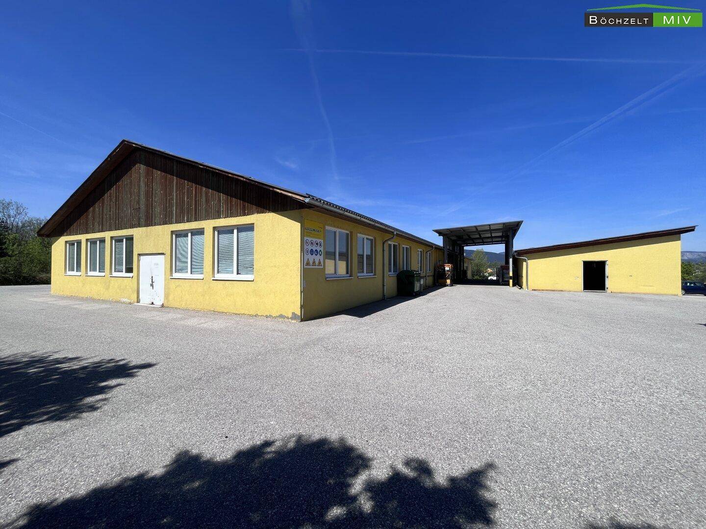 Gelbes Gewerbegebäude mit Holzgibel und großem Vorplatz unter strahlend blauem Himmel.