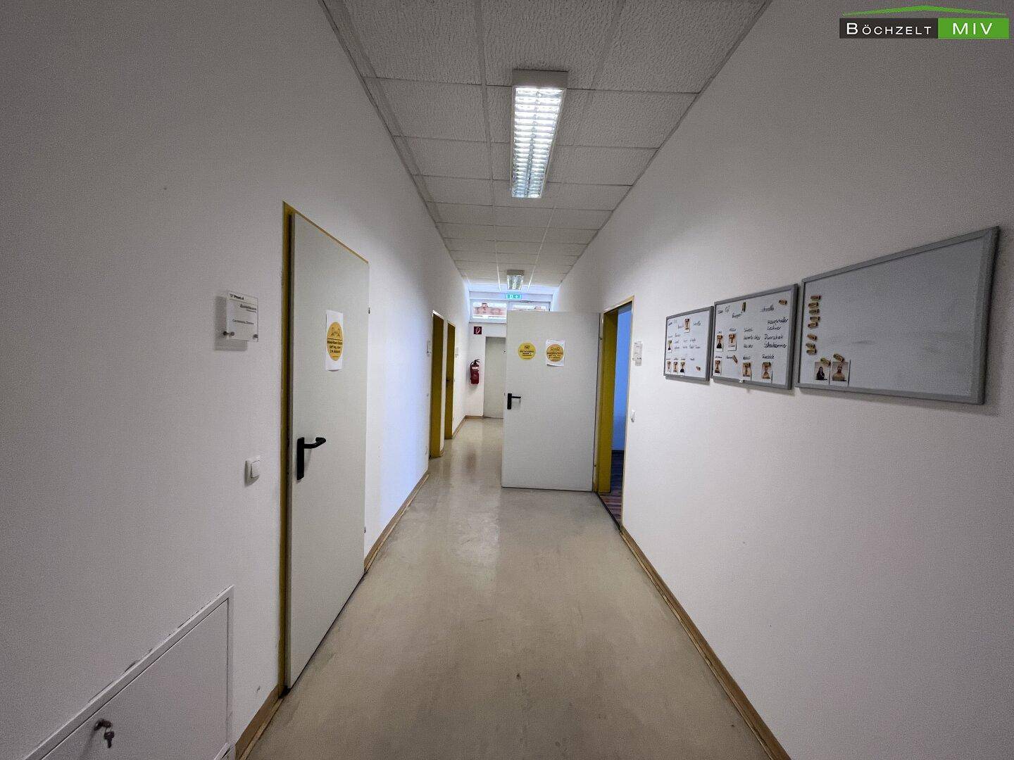 Langer, heller Flur mit weißen Wänden und mehreren Türen, ausgestattet mit Informationsaushängen und Beleuchtung.