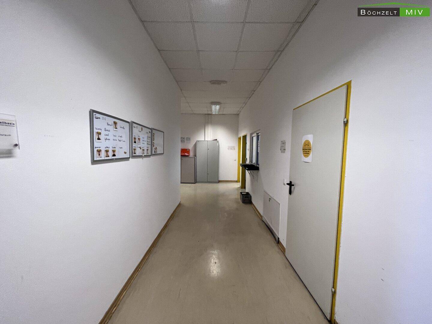 Heller Flur mit weißen Wänden und Türen, der zu verschiedenen Räumen führt, mit Informationsaushängen und Beleuchtung.
