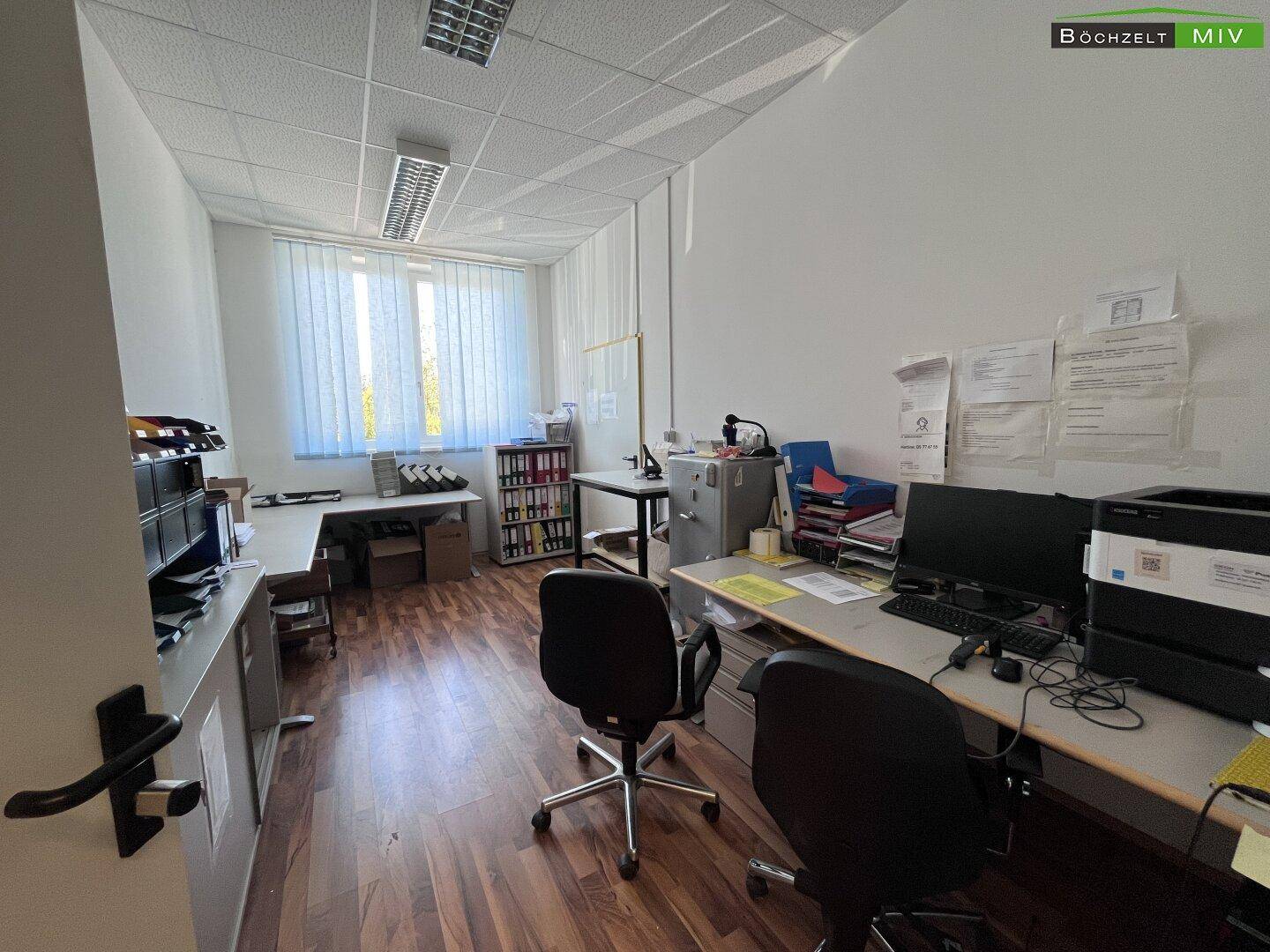 Helles Büro mit zwei Arbeitsplätzen, Schreibtischen und Regalen, gut beleuchtet durch große Fenster.