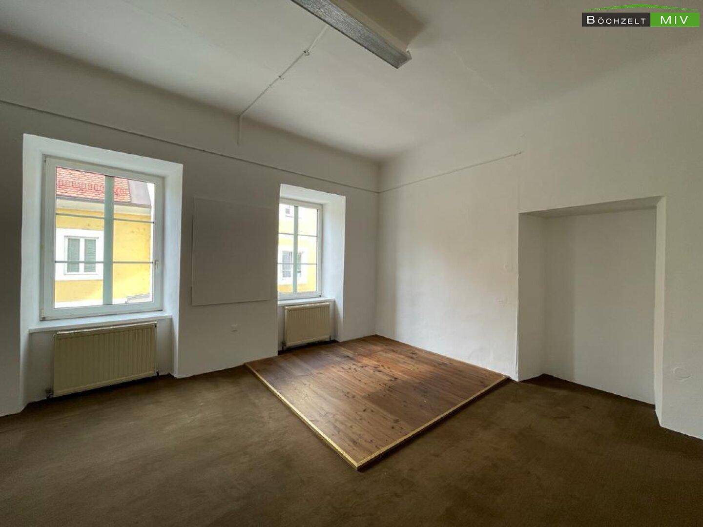 Obergeschossraum mit Teppichboden, Fenstern und einer erhöhten Holzplattform, vielseitig nutzbar.