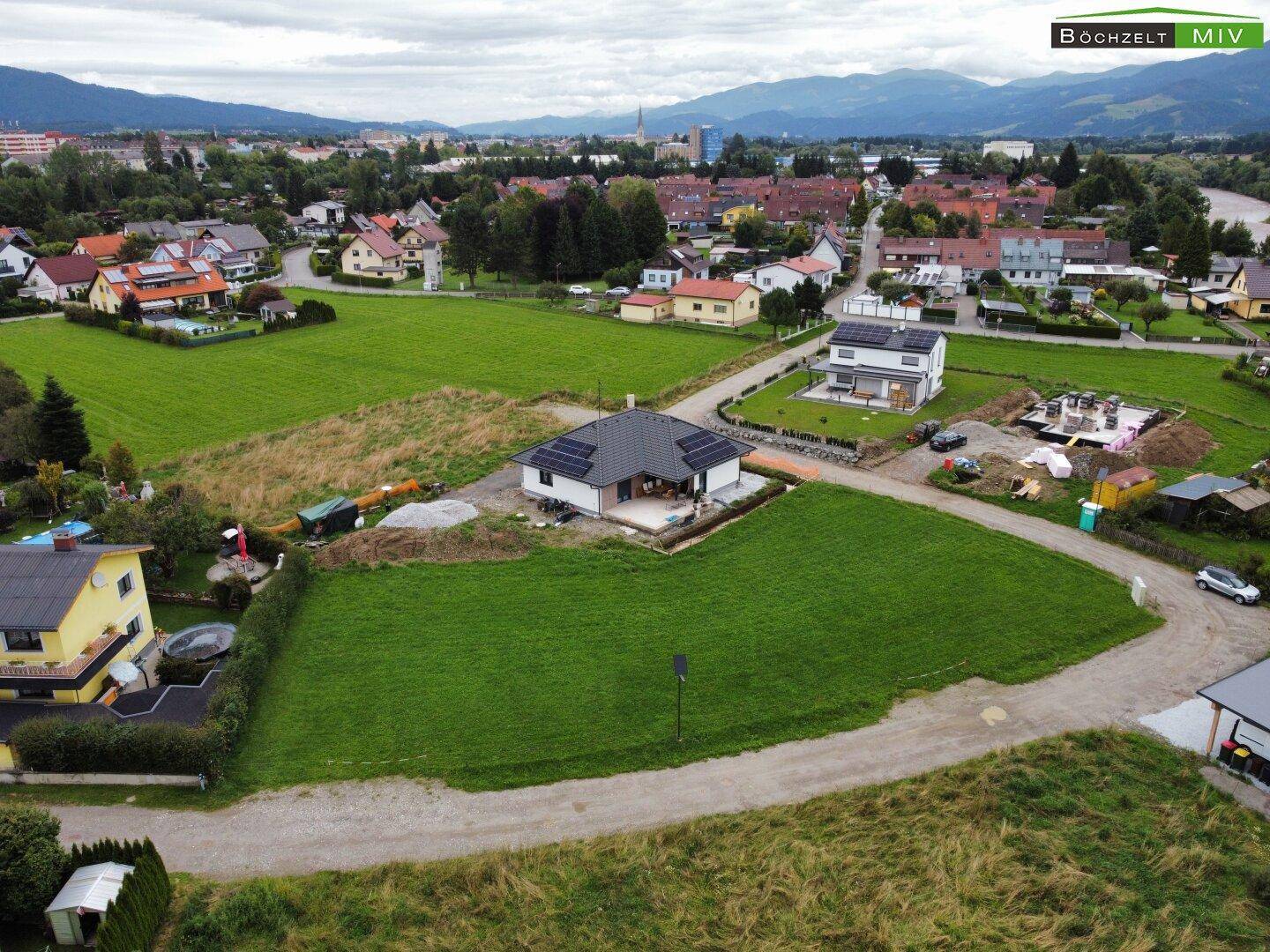 Luftaufnahme eines modernen Neubaus mit Solaranlage auf dem Dach, umgeben von Grünflächen und Bergen.