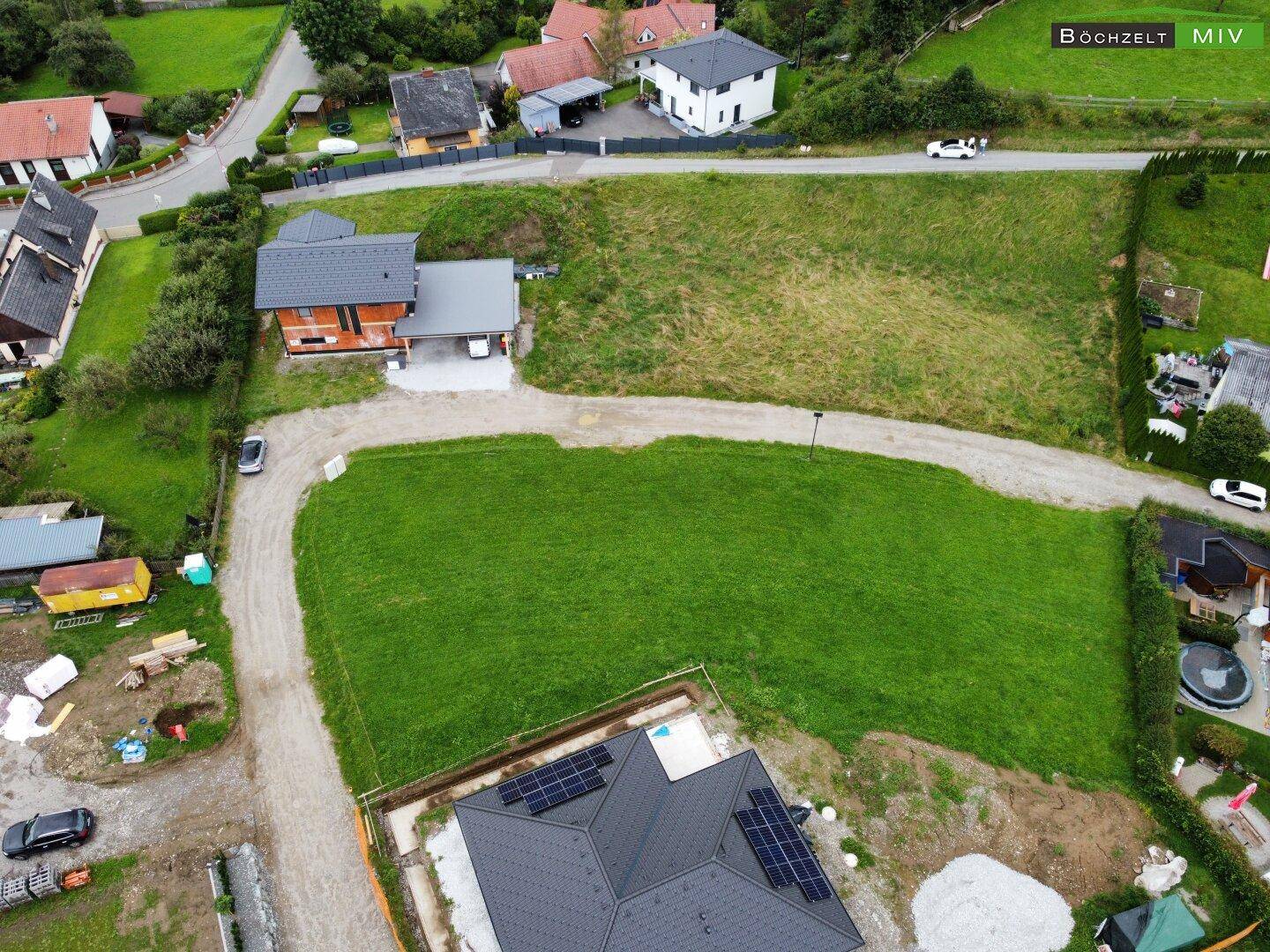 Blick von oben auf ein im Bau befindliches Haus mit Solaranlage und weitere Wohnhäuser in grüner Umgebung.