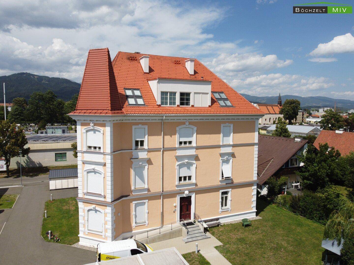Vorderansicht eines historischen Gebäudes mit beiger Fassade, roten Dachziegeln und vielen Fenstern.