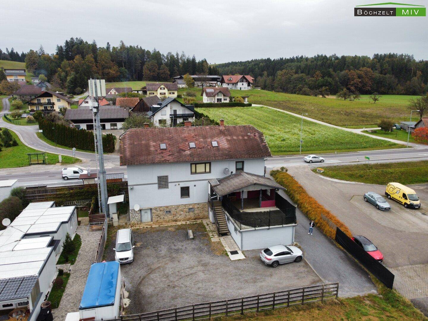 Luftaufnahme des Gebäudes von hinten mit Parkplätzen und angrenzender Straße, umgeben von grüner Landschaft.