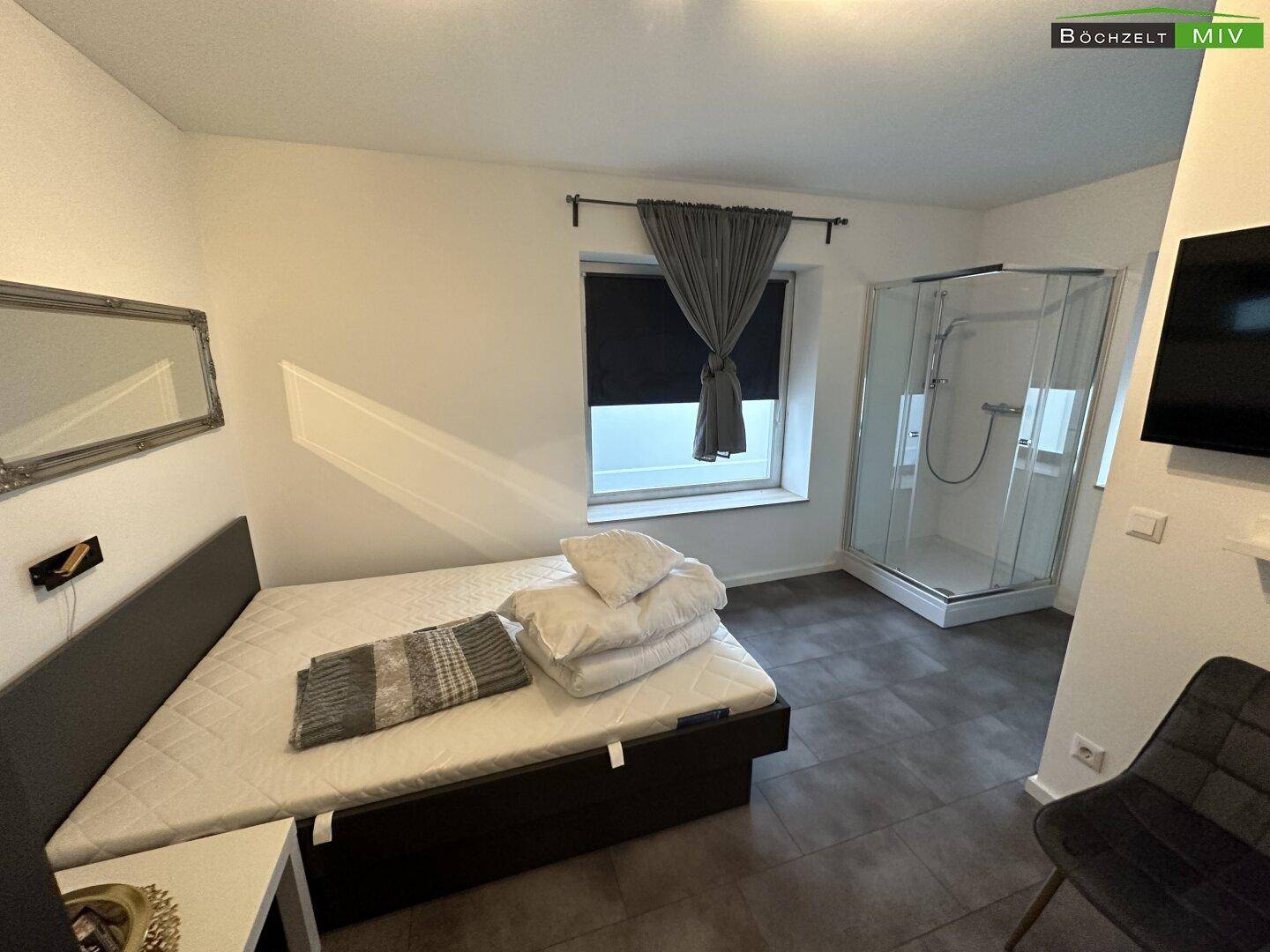 Schlafzimmer mit Bett, Fenster und einer integrierten Duschkabine im Raum, ausgestattet mit dunklen Vorhängen.