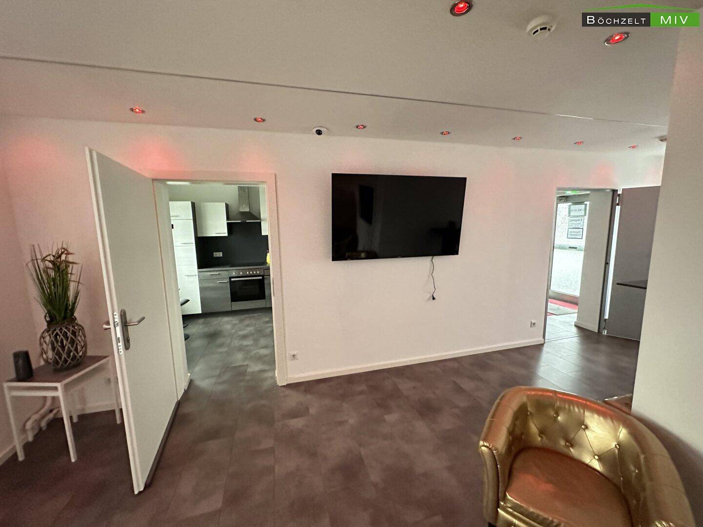 Wohnbereich mit Wandfernseher, Zugang zur Küche und einem goldenen Sessel, ausgestattet mit Überwachungskamera.
