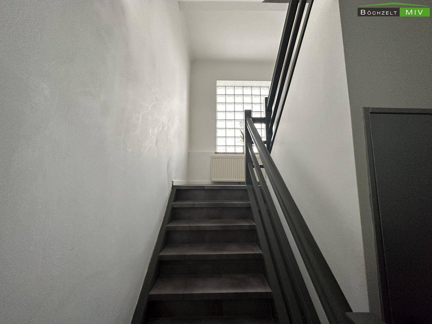 Treppenhaus mit dunklen Stufen und Geländer, beleuchtet durch ein Glasbausteinfenster, das Tageslicht hereinlässt.