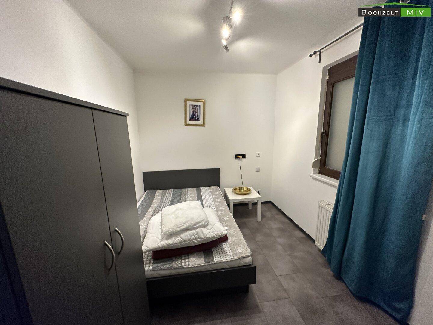Kleines Schlafzimmer mit Einzelbett, Kleiderschrank und Fenster mit blauem Vorhang, schlicht eingerichtet.