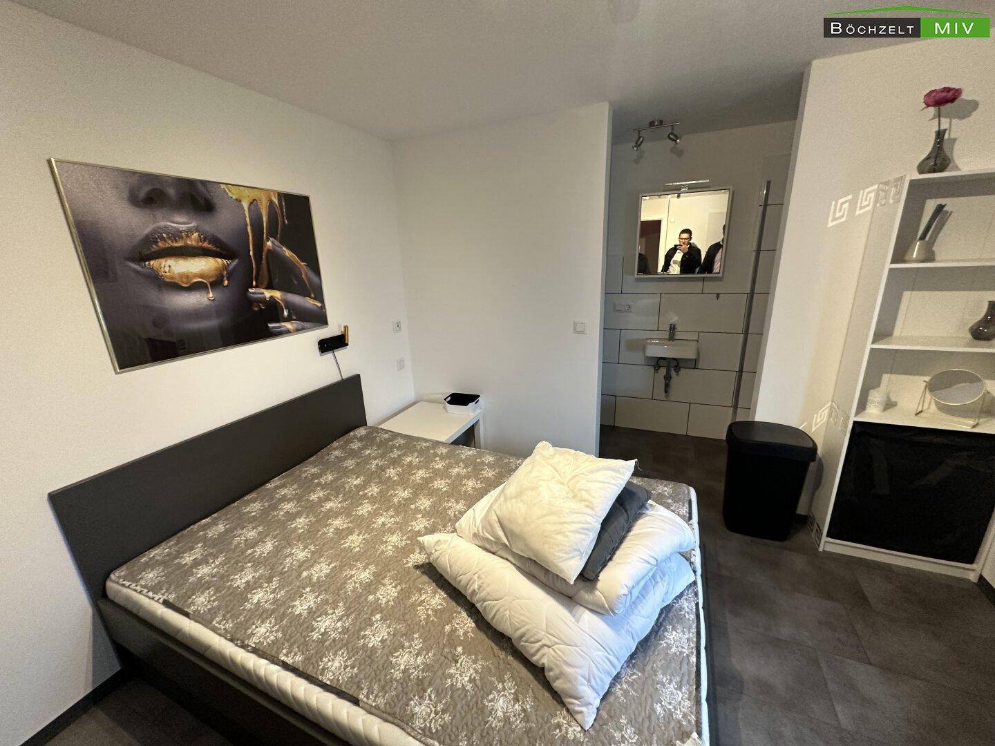 Schlafzimmer mit Bett, Wandbild und einem integrierten Badezimmerbereich mit Waschbecken und Spiegelschrank.