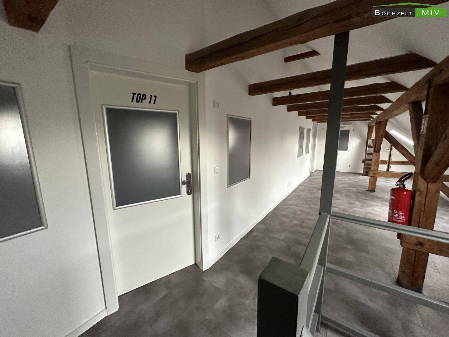 Dachgeschossflur mit sichtbaren Holzbalken, dunklem Boden und Zugang zu den Zimmern, hell gestrichen.