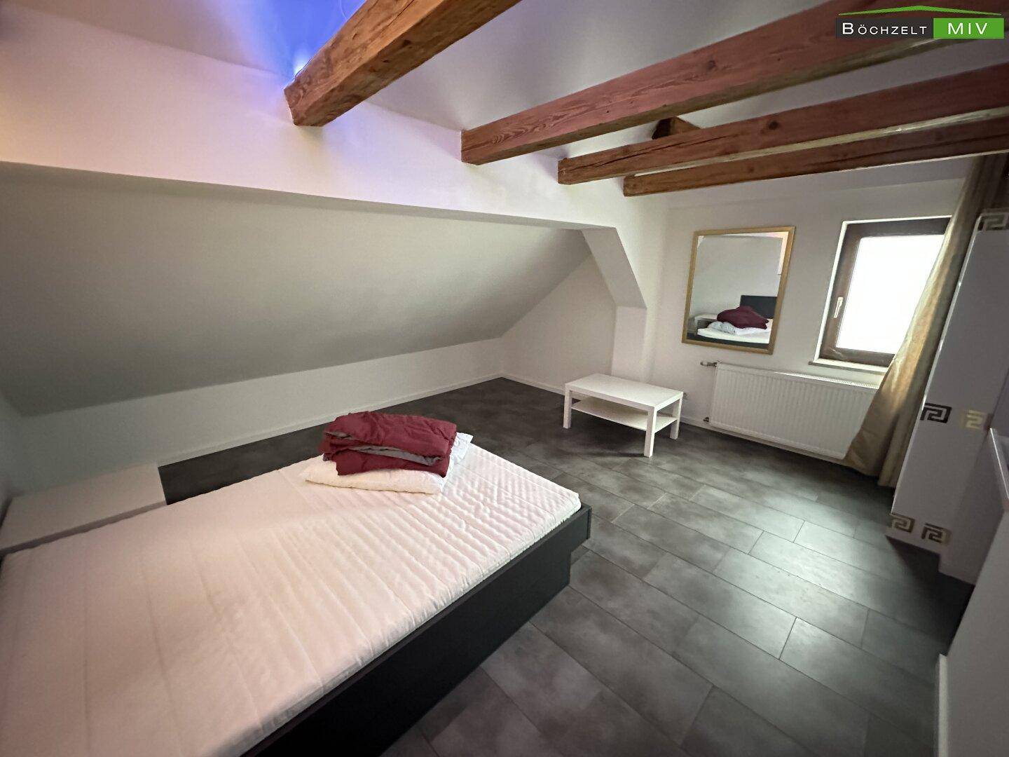 Dachgeschoss-Schlafzimmer mit Bett, Fenster, Heizkörper und sichtbaren Holzbalken, gemütlich eingerichtet.