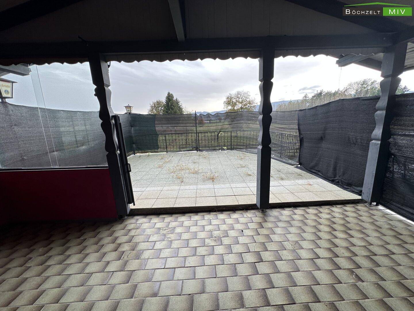 Überdachte Terrasse mit Fliesenboden und weitem Blick auf die umliegende grüne Landschaft unter bewölktem Himmel.
