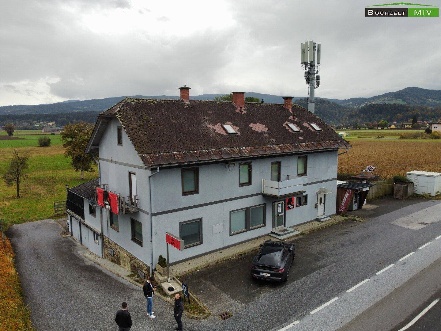 Luftaufnahme des Gebäudes mit umliegender Landschaft, Feldern und einem Mobilfunkmast unter bewölktem Himmel.