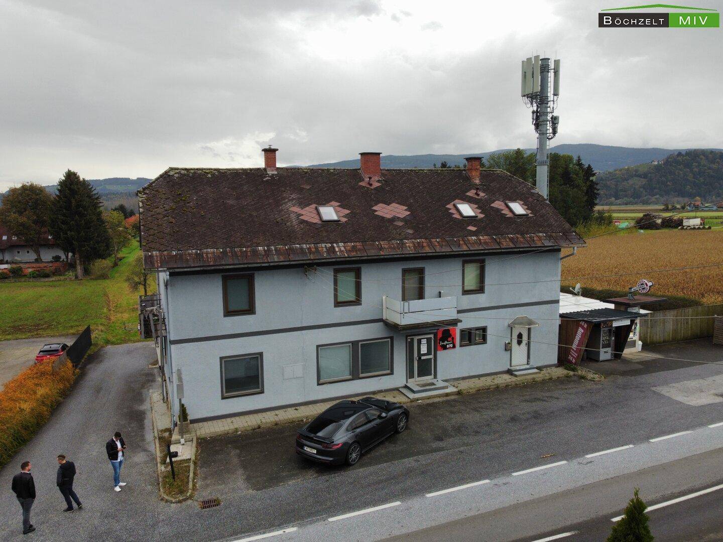 Luftaufnahme des Gebäudes mit umliegender Landschaft, Feldern und einem Mobilfunkmast unter bewölktem Himmel.