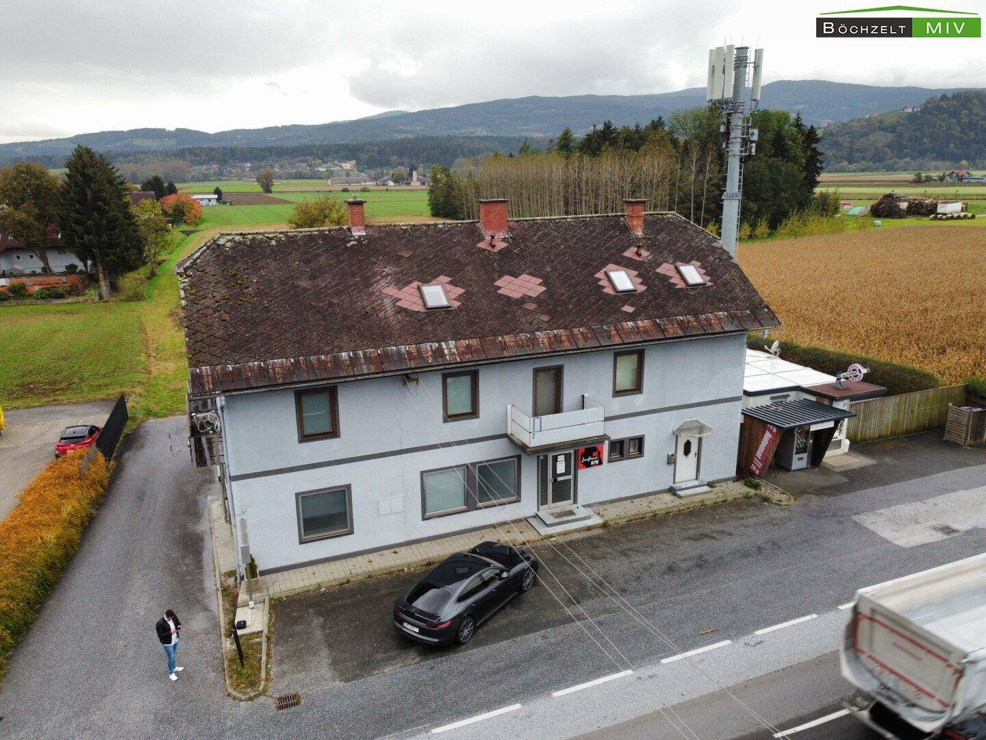 Luftaufnahme des Gebäudes mit umliegender Landschaft, Feldern und einem Mobilfunkmast unter bewölktem Himmel.