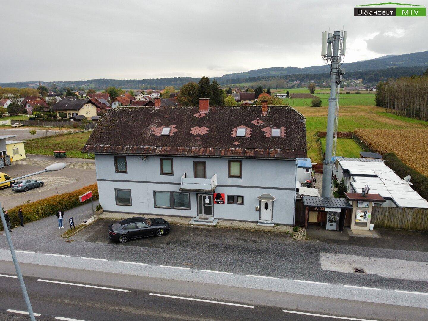 Luftaufnahme des Gebäudes mit umliegender Landschaft, Feldern und einem Mobilfunkmast unter bewölktem Himmel.