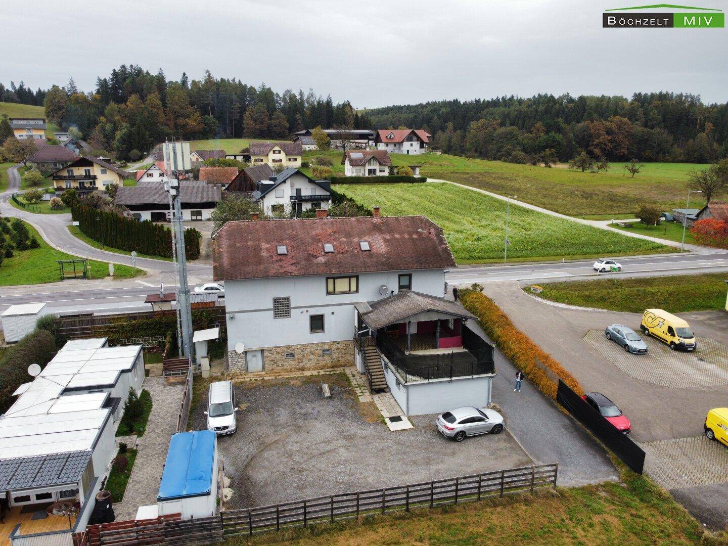 Luftaufnahme des Gebäudes von hinten mit Parkplätzen und angrenzender Straße, umgeben von grüner Landschaft.