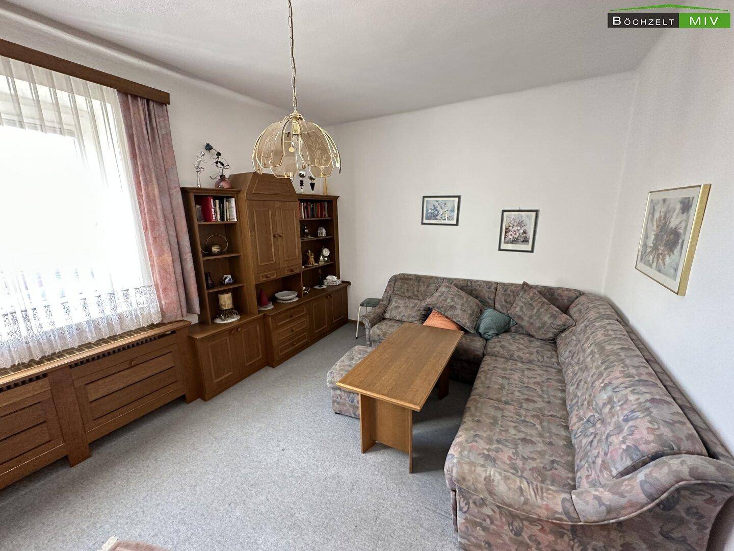 Wohnzimmer mit großem Sofa, Holzwandmöbeln und Teppichboden, hell durch ein großes Fenster.