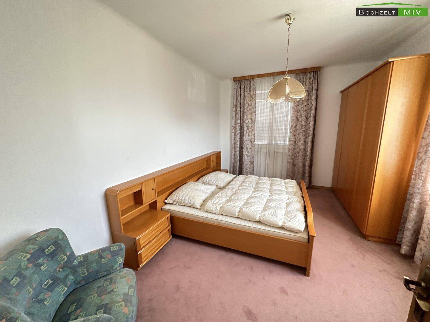 Schlafzimmer mit Doppelbett, Holzschrank und Teppichboden, hell durch zwei Fenster.